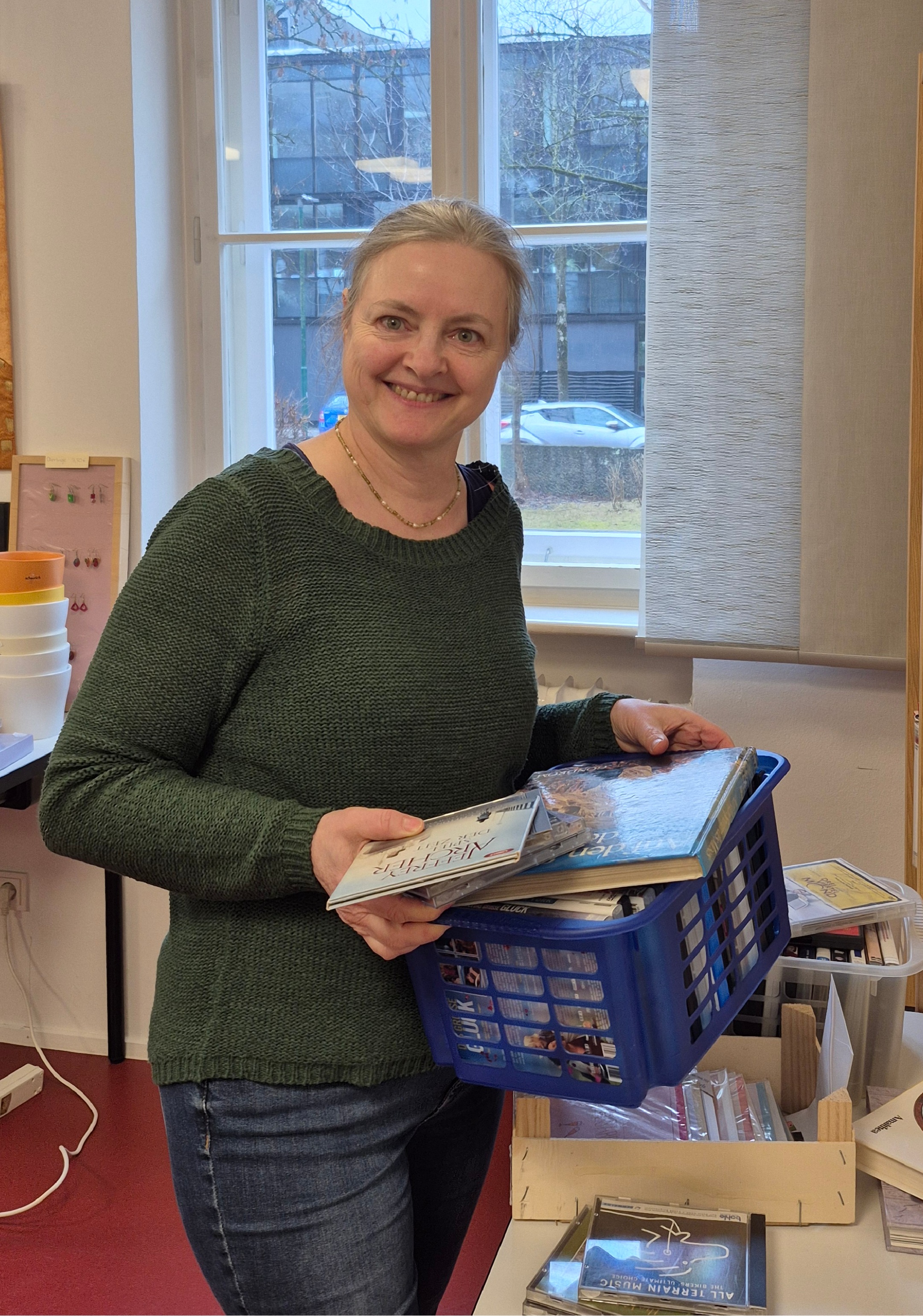 Antje Lohrer lächelt in die Kamera, in den Händen eine Kiste voller CDs, DVDs und Bücher, die sie gerade sortiert.